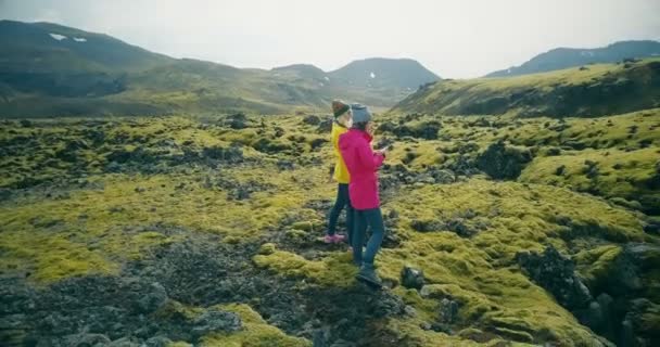 Luftaufnahme von zwei Touristinnen, die auf Lavafeldern in Island spazieren und Fotos machen. Mädchen nutzen Smartphone und Kamera. — Stockvideo