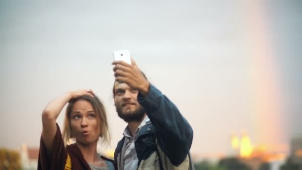 Casal jovem tirando fotos selfie com arco-íris no fundo. Homem e mulher alegres usam a tecnologia do smartphone . — Vídeo de Stock