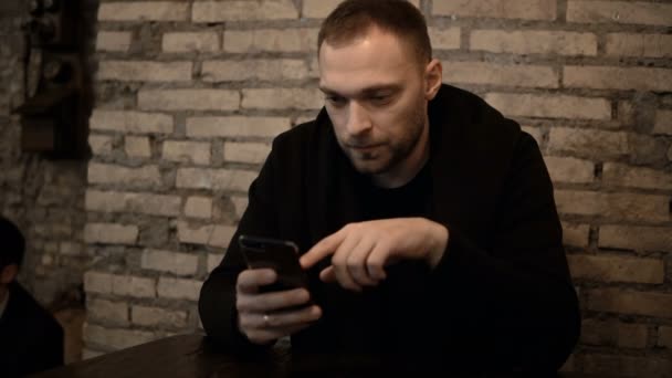 Young handsome man sitting in the cafe with brick wall and using the smartphone, browsing the Internet alone. — Stock Video