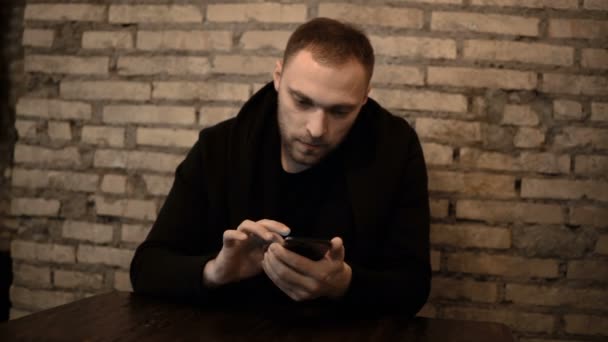 Jovem homem bonito sentado no café perto da parede de tijolo e usando o smartphone, navegando na Internet sozinho . — Vídeo de Stock
