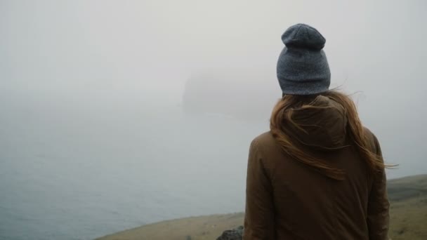 Back view of young woman in hat standing on the shore of the sea and thinking about something, exploring the Iceland. — Stock Video