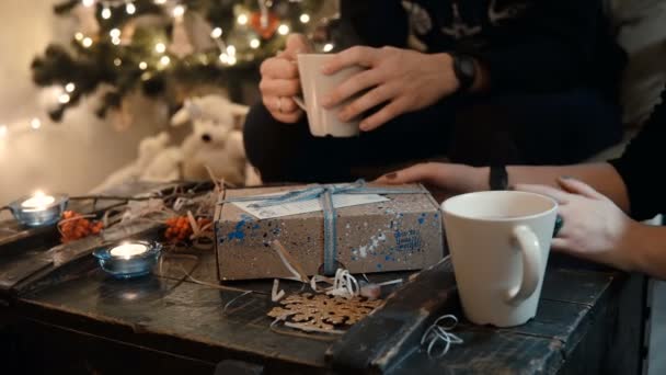 Close-up beeld van jonge paar zitten aan de tafel, in de buurt van de kerstboom en open de vakantie aanwezig, het drinken van thee. — Stockvideo