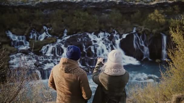 Baksidan på ung resande par stående i bergen nära vattenfallen på Island och ta foto på smartphone — Stockvideo