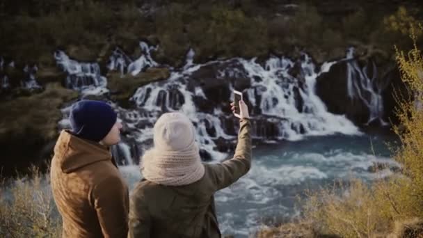 Mladý cestující pár stojící v horách údolí nedaleko vodopádu a mluvící obrázky nebo fotografie selfie na smartphone. — Stock video