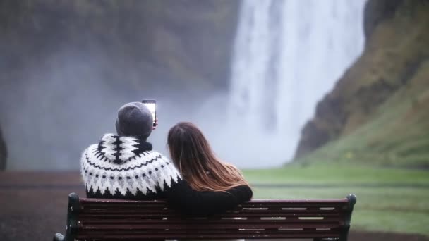 Baksidan på unga paret sitter på bänken och ta selfie foto på smartphone nära vattenfallet Skogafoss i Island — Stockvideo