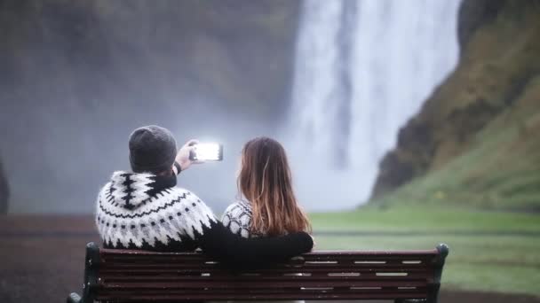 Ungt par tar fotot på smartphone av det vackra vattenfallet Skogafoss i Island, berömda turist plats. — Stockvideo