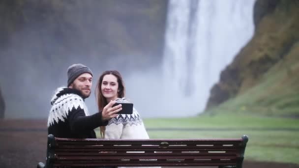 Młoda para szczęśliwy, siedząc na ławce i biorąc zdjęcie na smartfon w pobliżu wodospadu Skogafoss w Islandii. — Wideo stockowe