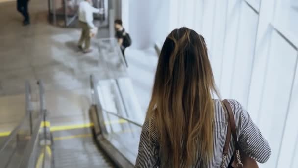 Vue arrière de la jeune belle femme debout sur descendre l'escalator et en utilisant le smartphone, se préparant à la réunion . — Video