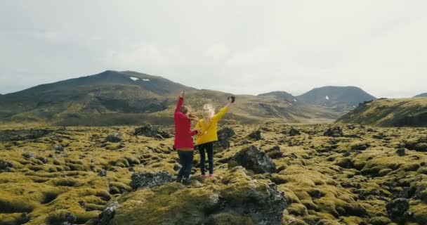 Vista aérea de dos mujeres de pie sobre la roca en el campo de lava en Islandia. Los turistas miran el paisaje, bailan y saltan — Vídeos de Stock