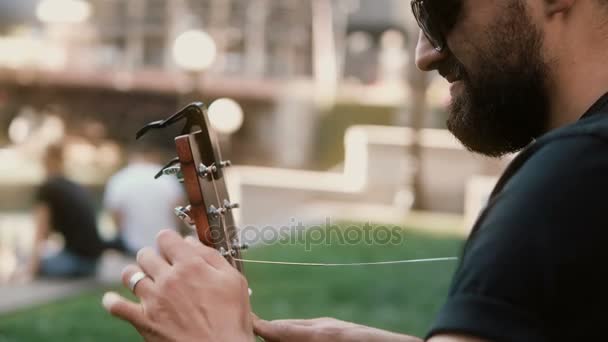 Vista da vicino del giovane in occhiali da sole trattenuto, cambiando corda in chitarra per strada nella soleggiata giornata estiva . — Video Stock