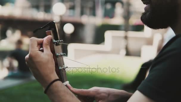 Närbild av ung stilig man med skägg restrung, ändra strängen i gitarr på gatan i solig sommardag. — Stockvideo