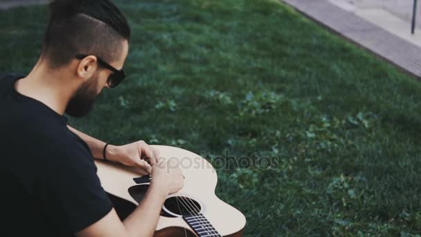 Joven hombre guapo con barba sentado en el césped en el parque y cambiando cuerdas, restringió la guitarra fuera . — Vídeos de Stock