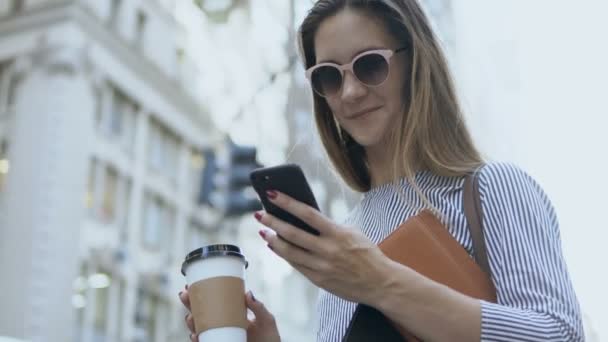 Porträt einer jungen schönen Geschäftsfrau mit Sonnenbrille, die die Tasse Kaffee mit dem Smartphone auf der Straße hält. — Stockvideo