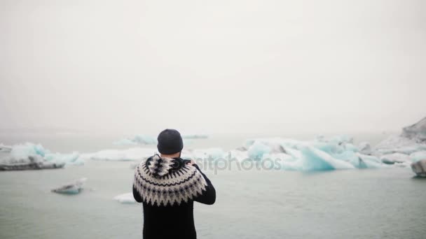 Back view of young traveling man standing in Jokulsalon ice lagoon in Iceland and take photos og glaciers on smartphone. — Stock Video