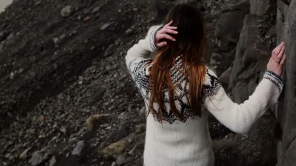 Vista posterior de la joven morena en lopapeysa caminando a través de las rocas, senderismo en las montañas solo en el día ventoso . — Vídeo de stock