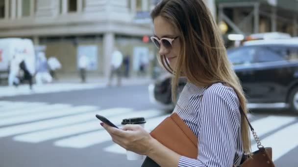 Young beautiful businesswoman holding the documents and coffee cup. Busy female using smartphone standing near the road. — Stock Video