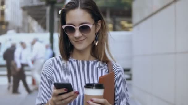 Joven mujer de negocios feliz caminando por la calle, sosteniendo taza de café y documentos, utilizando el teléfono inteligente . — Vídeos de Stock