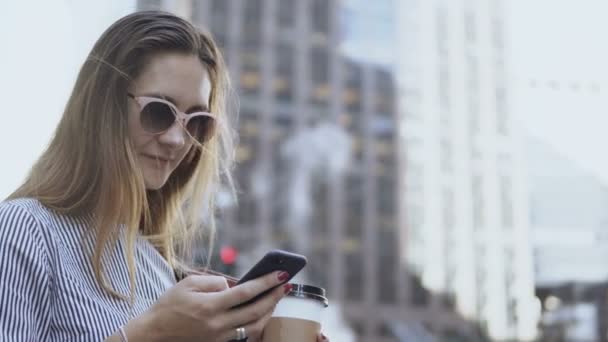 Portret van jonge zakenvrouw in zonnebril met behulp van de smartphone, houdt het koffiekopje op straat tijdens pauze. — Stockvideo