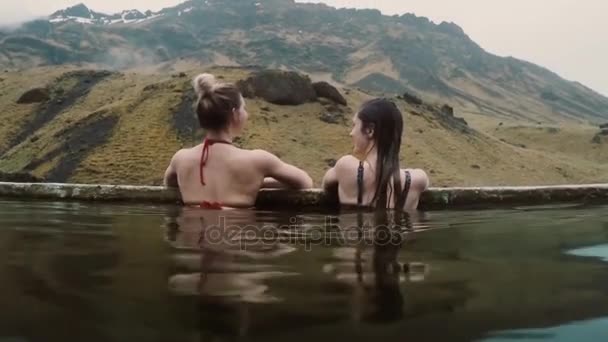 Termas geotermales en Islandia. Dos mujeres viajeras relajándose en la piscina caliente con hermoso paisaje en la montaña . — Vídeos de Stock