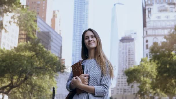 Retrato de una joven mujer de negocios feliz de pie en el distrito financiero de Nueva York, cerca de rascacielos y sostiene cuadernos . — Vídeos de Stock