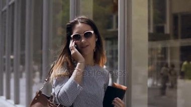 Belgeleri holding ve sokakta yürürken genç iş kadını. Mutlu kadın cep telefonu üzerinde konuşurken. Ağır çekim.