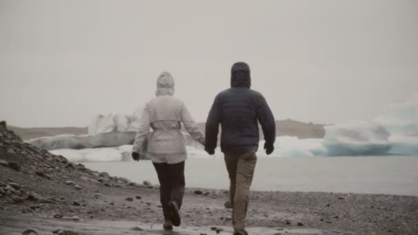 Vista posterior de una joven pareja caminando en la laguna de hielo en Islandia. Hombre y mujer explorando los icebergs y glaciares . — Vídeos de Stock