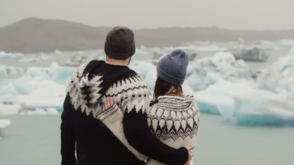 Вид сзади на молодую пару, стоящую в ледяной лагуне. Мужчина целует женщину и обнимает ее, глядя на ледники в Исландии . — стоковое видео