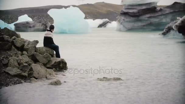 Joven hombre guapo sentado en la roca en la laguna de hielo y disfrutando de la vista. Hombre explorando la Islandia solo . — Vídeos de Stock