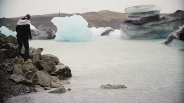 Joven hombre guapo caminando en la laguna de hielo. Felices turistas se sientan en la roca y observan los glaciares en Islandia . — Vídeo de stock