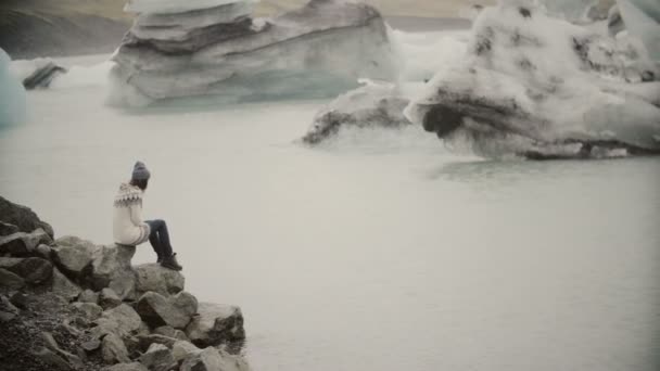 Junge attraktive Frau, die auf einem Felsen in der Eislagune sitzt. Tourist in lopapeysa erkundet die Aussicht auf Island. — Stockvideo
