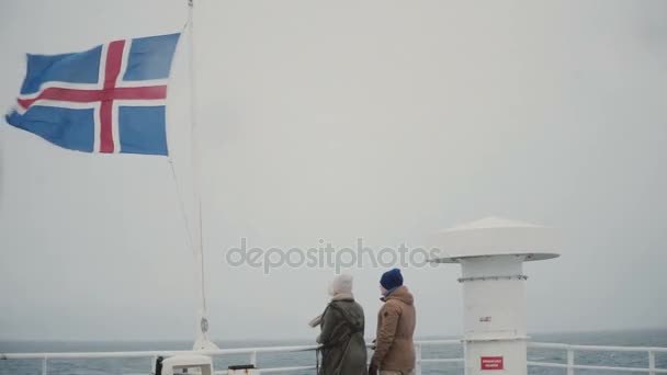 Vista trasera de la joven pareja de pie a bordo del barco, cerca de la bandera de Islandia y mirando al mar, hablando . — Vídeo de stock