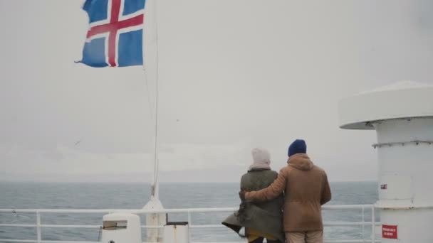 Vista trasera de una pareja joven y elegante de pie a bordo del barco con bandera islandesa. Hombre y mujer miran al mar . — Vídeo de stock