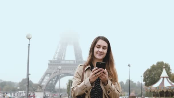 Ritratto di giovane bella donna in piedi in mattinata nebbiosa vicino alla torre Eiffel a Parigi, Francia e utilizzando lo smartphone . — Video Stock