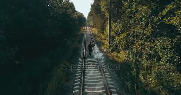 Homem a correr em carris de comboio com uma bomba de fumo. Visão traseira do drone. Corredor sinalizando para o trem que vem. Ferrovia florestal de outono . — Vídeo de Stock