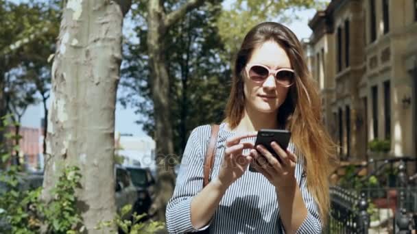 Retrato de una joven hermosa mujer en gafas de sol caminando afuera en un día soleado de verano y usando un teléfono inteligente. Movimiento lento . — Vídeos de Stock