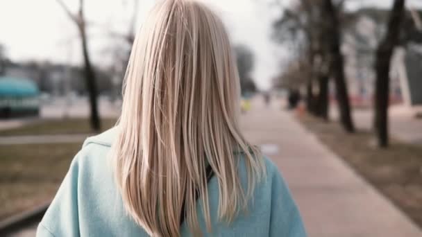 Блондинка с видом сзади ходит и смотрит влево. Белая девушка в синем пальто с красивыми длинными светлыми волосами. Медленное движение . — стоковое видео