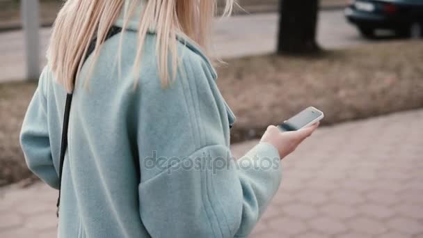 Rubia de cámara lenta adolescente en azul abrigo mensajes de texto. Vista lateral. Chica con el pelo largo de lectura desde el teléfono inteligente. Transmisión web . — Vídeos de Stock
