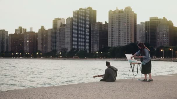 Joven pareja hermosa en la orilla del lago Michigan, Chicago, Estados Unidos. Mujer dibujar, hombre tocar la guitarra . — Vídeo de stock