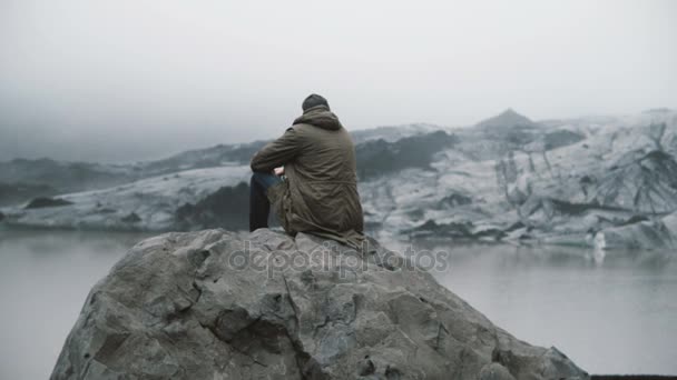 Zadní pohled na sání Asijské twink sedí na skále a při pohledu na ledovce v ledovou lagunu. Muž poznávání Islandu sám. — Stock video