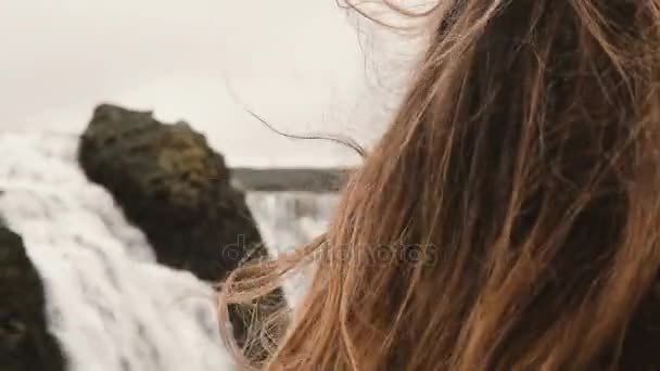 Portrait of young beautiful sad woman standing near the waterfall in Iceland and looking around. Hair waving on wind. — Stock Video