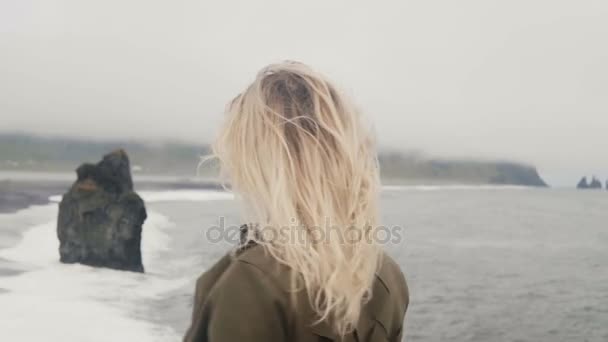 Молодая блондинка, стоящая на черном пляже в Айсленде, глядя на тролль пальцы камней, волосы машут по ветру . — стоковое видео