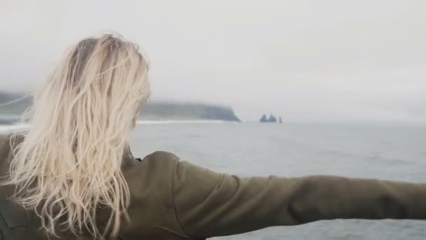 Вид на молодую блондинку, стоящую на черном пляже в Айсленде. Свобода: турист поднимает руки, волосы машут на ветру — стоковое видео