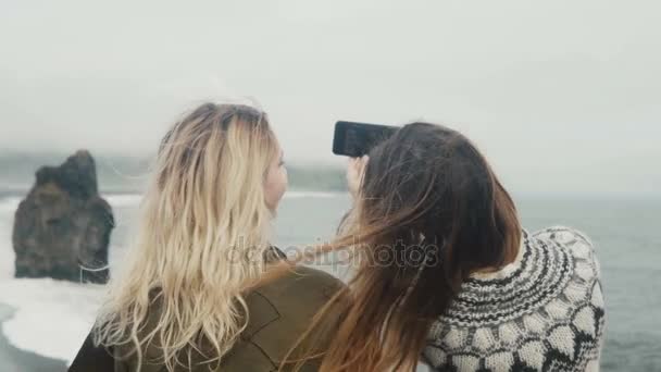 Vista posterior de dos mujeres jóvenes tomando fotos selfie en la orilla del mar cerca de las rocas de los dedos de los pies troll en Islandia . — Vídeos de Stock