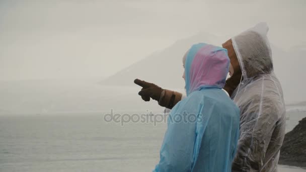 Jeune couple en imperméable debout dans la lagune de glace en Islande et montrant quelque chose les uns aux autres avec les doigts . — Video