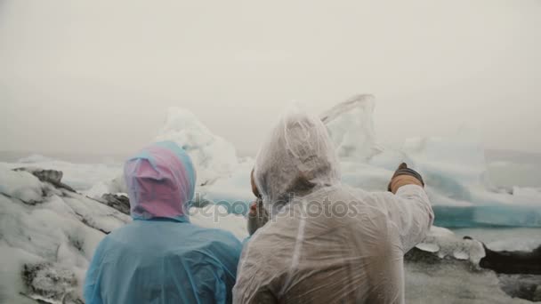 Back view of young couple in raincoats standing in ice lagoon in Iceland and looking on glaciers with ash, famous sight. — Stock Video