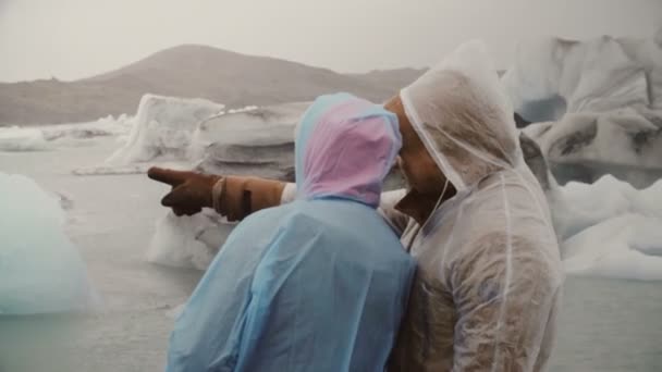 Junges Paar im Regenmantel beim Erkunden der berühmten Sehenswürdigkeit - der Eislagune in Island. Touristenmann zeigt Frau etwas. — Stockvideo