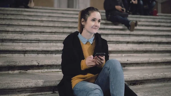 Estudante menina sentada à noite na escada e olhando para alguém, sorrir. Mulher usar smartphone, navegar na Internet . — Fotografia de Stock