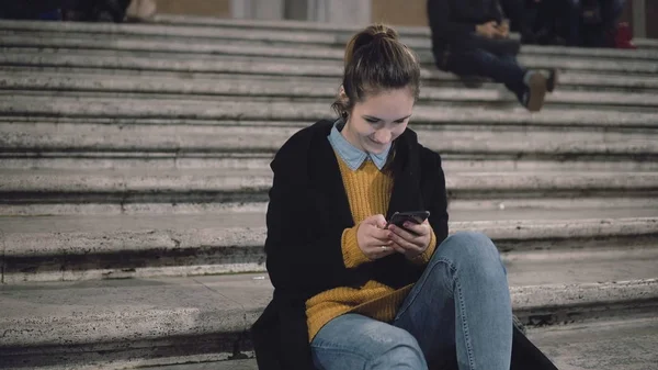 Jovem mulher atraente sentado na escada na cidade e usar smartphone. Estudante menina mensagens com amigos . — Fotografia de Stock