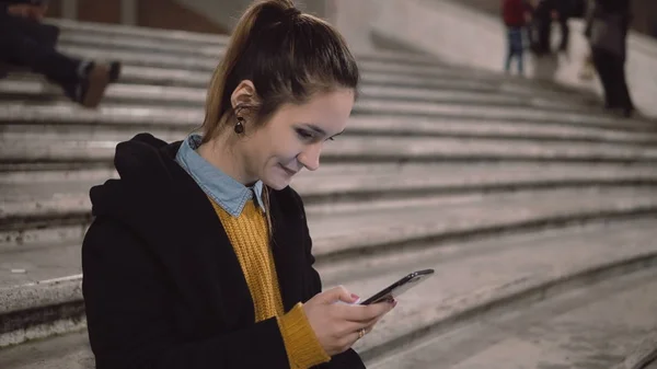 Retrato de jovem mulher sentada na escada e segurando smartphone. Menina usa touchscreen para navegar na Internet . — Fotografia de Stock