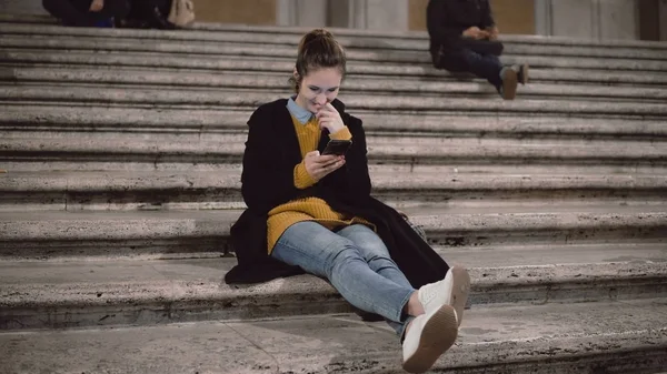 Attraktiv ung student sitter på stege och med smartphone. Flicka chattar med touchscreen-teknik — Stockfoto